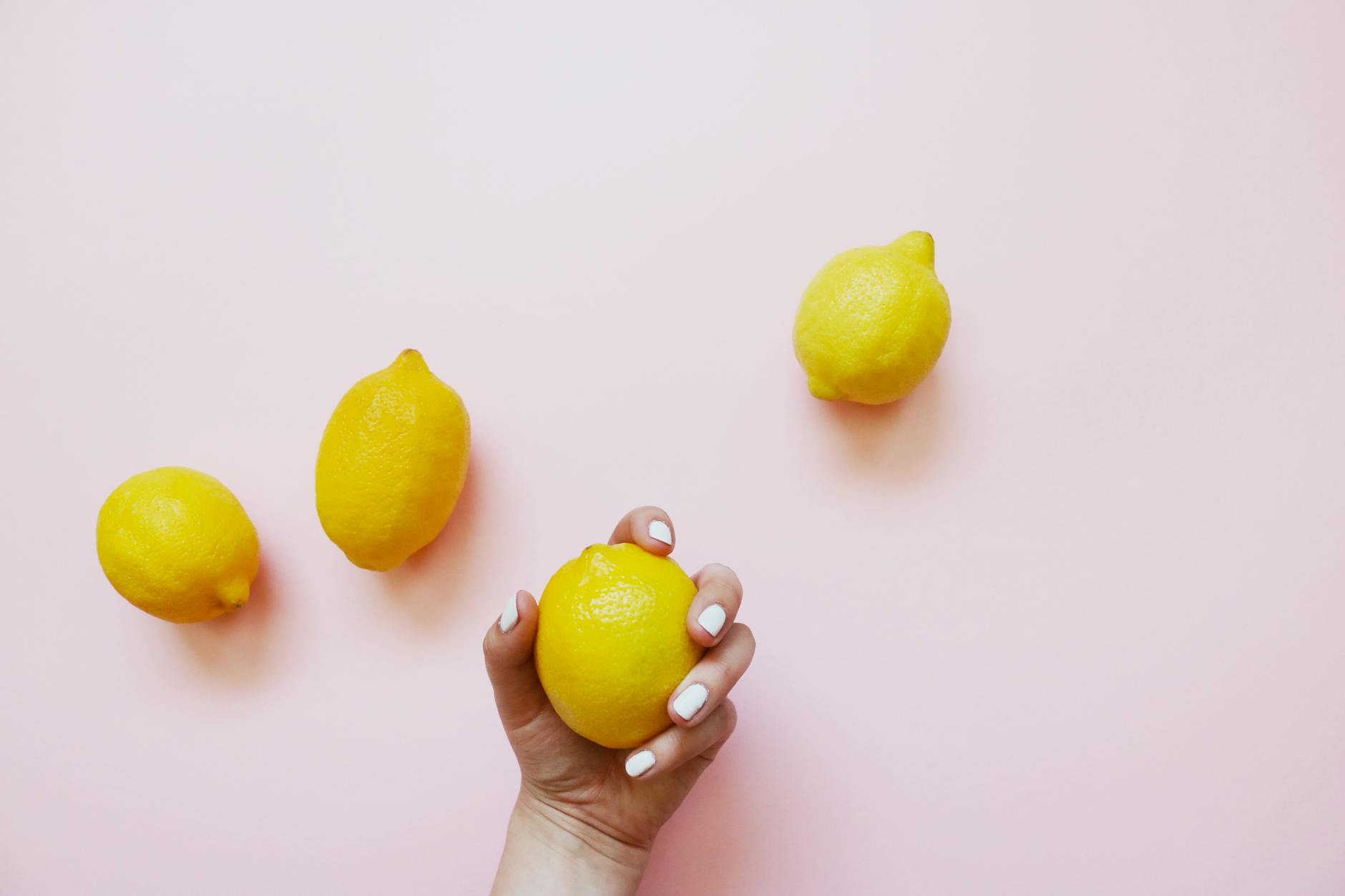 A hand with white nails holding a fresh lemon on a soft pink background, surrounded by additional lemons